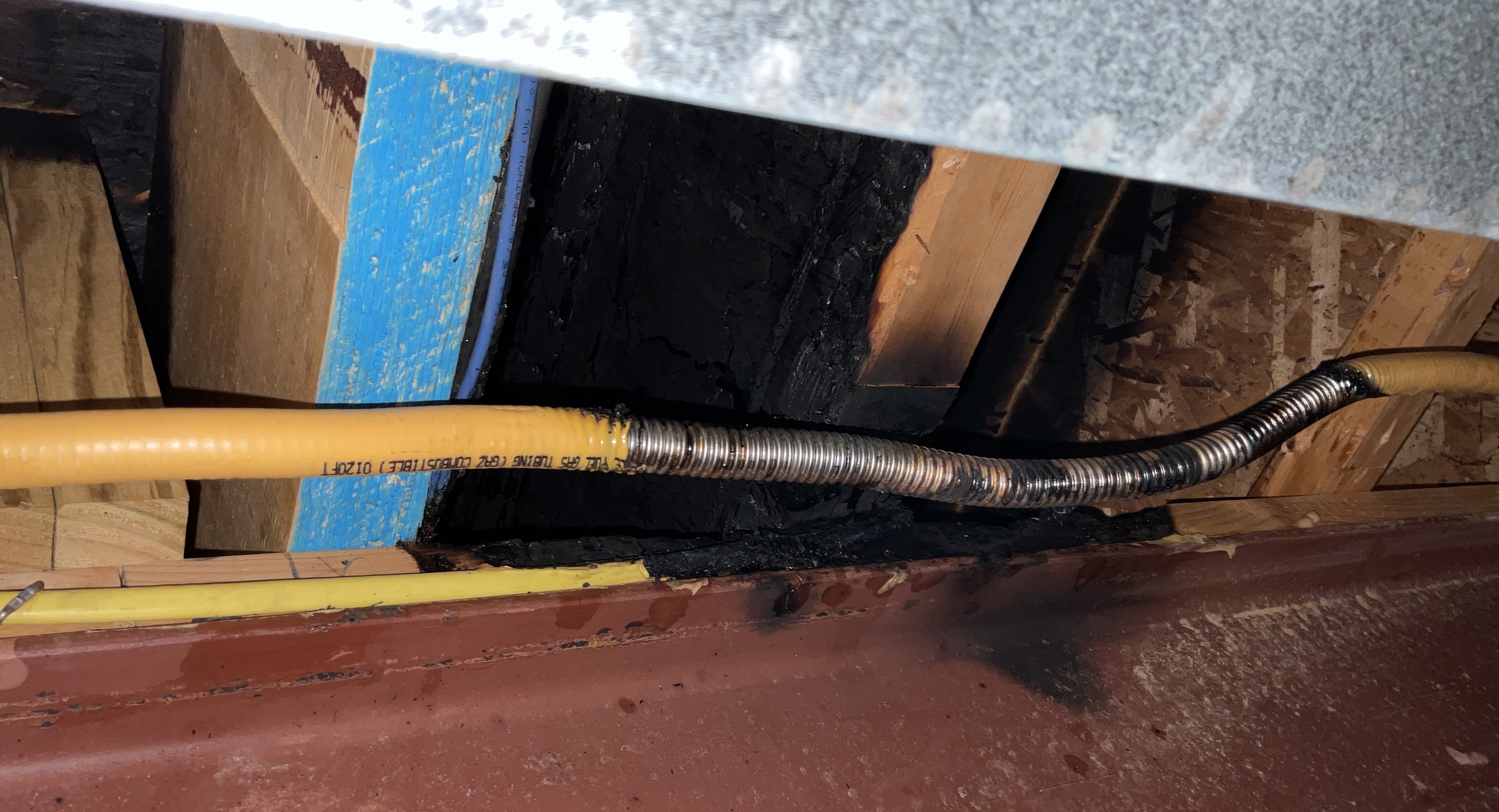 A charred seciton of ceiling joist sits right above a 
section of corrugated stainless steel gas line. At either end, it is covered with yellow 
plastic, but that's melted off this section. It's near a red-painted steel I-beam;
 a small section of galvanized rectangular heating-cooling duct is visible nearby.